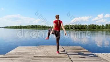 美丽的，运动的年轻金发女人伸展，做不同的运动，跳跃，跳跃，蹲。 湖泊，河流，蓝色