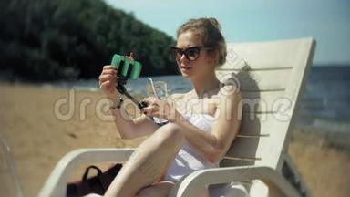 一个穿着白色比基尼的年轻女孩躺在沙滩上的躺椅上晒太阳，还在智能手机上拍照