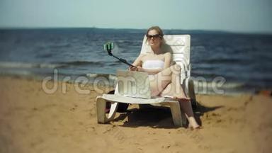 一个穿着白色比基尼的年轻女孩躺在沙滩上的躺椅上晒太阳，还在智能手机上拍照