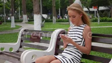 年轻漂亮的苗条女人，一头金发，穿着<strong>黑白</strong>连衣裙，坐在长凳上，用智能<strong>手机</strong>上网