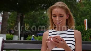 年轻漂亮的苗条女人，一头金发，穿着黑白连衣裙，坐在长凳上，用智能手机上网