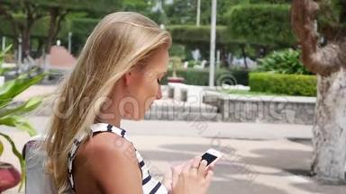 年轻漂亮的苗条女人，一头金发，穿着黑白连衣裙，坐在长凳上，用智能手机上网
