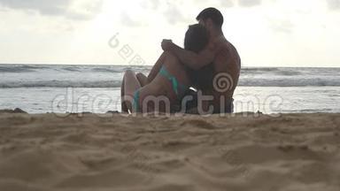 年轻的浪漫夫妇正在享受美丽的景色，坐在海滩上，晚上拥抱。 一个女人一个男人坐着