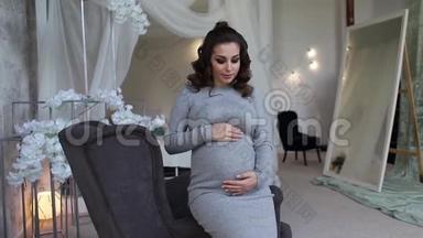 美丽的孕妇坐在扶手椅上，双手放在肚子上。 女人穿着优雅的衣服。 美丽美丽