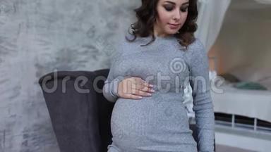 美丽的孕妇坐在扶手椅上，双手放在肚子上。 女人穿着优雅的衣服。 美丽美丽