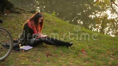 美丽的白种人微笑的女孩骑着<strong>自行车</strong>坐在公园里，<strong>学生</strong>在湖边的笔记本上写着，温暖