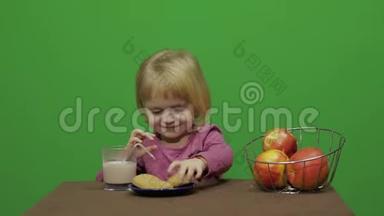 女孩坐在桌子旁吃巧克力、饼干和饮料可可。 快乐的三岁女孩。 可爱的女孩微笑