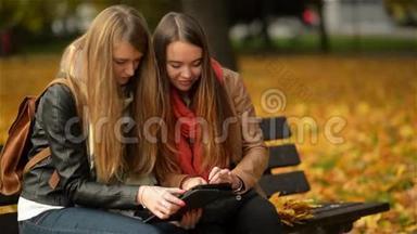 两个有趣的快乐年轻女孩朋友坐在长凳上用平板电脑。 女人在Pad电脑上嬉笑打闹