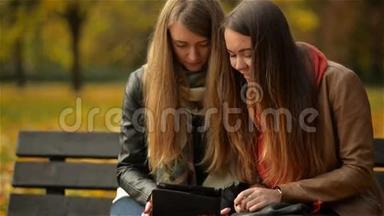 两个有趣的快乐年轻女孩朋友坐在长凳上用平板电脑。 女人在Pad电脑上嬉笑打闹