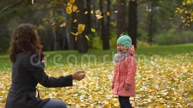 快乐的年轻妈妈和她的小女儿在秋天的公园里玩，妈妈和女孩扔树叶和笑。 慢慢慢慢