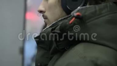 一位身穿夹克衫、戴着大耳机的长胡子青年坐在电车旁的公共交通工具里