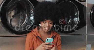 在<strong>洗衣店</strong>用智能手机微笑的年轻女人。 自助公共<strong>洗衣店</strong>。