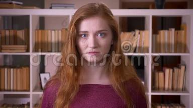 在大学图书馆里，一位年轻的红头发、迷人的白种人女学生看着摄像机的特写镜头