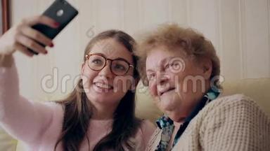 奶奶和年轻女孩在家里坐在沙发或沙发上用手机或智能手机拍照。 快乐的女士们