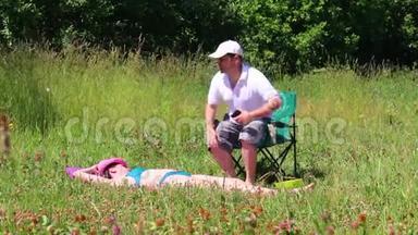 一个穿泳衣的女孩正在草坪上晒日光浴。 在她旁边的野<strong>餐椅</strong>上，一个男人坐在智能手机里玩。