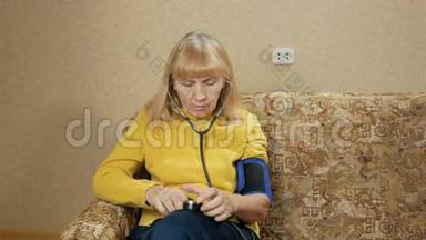 老年妇女在沙发上测量自己家的压力。 她通过血压计听脉搏