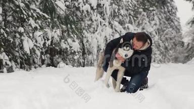 一个男人在森林里拥抱他的西伯利亚哈士奇。 在雪地里的背景树上。 眼中的骄傲和幸福.. 步行