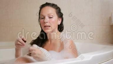 一个年轻的女孩用泡沫洗澡，用洗发水洗头。 一个大的白色浴缸和一小瓶洗发水