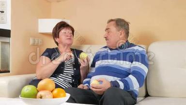 老两口在家里躺在沙发上吃苹果。 桌子上有一盘不同的水果。 丈夫和