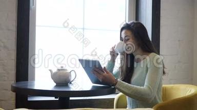 年轻的黑发女孩坐在靠近窗户的桌子上，看着平板电脑。 喝茶。
