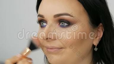 造型师化妆师用一种特殊的刷子在年轻的蓝色美女的脸上涂上粉底