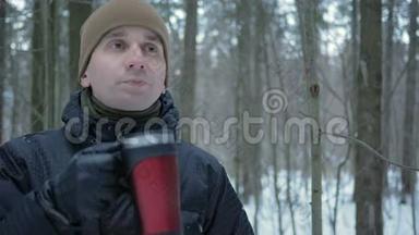 一个年轻人在冬天的森林里<strong>喝热水</strong>瓶里的热饮料。 那个人看着镜头，穿着深色衣服