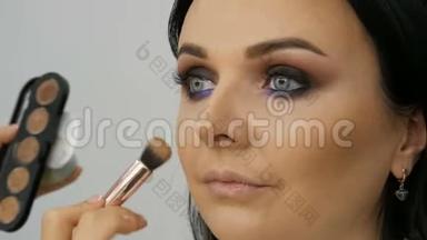 造型师化妆师用一种特殊的刷子在年轻的蓝色美女的脸上涂上<strong>粉底</strong>