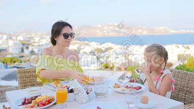 <strong>一家人</strong>在室外咖啡馆<strong>吃早餐</strong>，在Mykonos镇有令人惊叹的景色。 可爱的女孩和妈妈喝着新鲜果汁