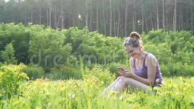 快乐的年轻女子坐在绿色的草坪上，在夕阳的背景下，在风景名胜区使用智能手机