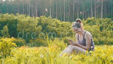 快乐的年轻女子坐在绿色的草坪上，在夕阳的背景下，在风景名胜区使用智能手机
