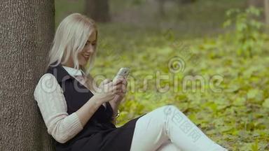 秋天公园里一个美丽的女孩坐在树下，手里拿<strong>着手机</strong>，微笑<strong>着看着</strong>留言