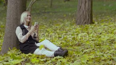 秋天公园里一个美丽的女孩坐在树下，手里拿着手机，微笑着看着留言