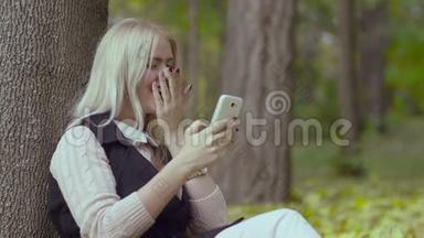 秋天公园里一个美丽的女孩<strong>坐在树下</strong>，手里拿着手机，微笑着看着留言