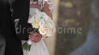 优雅的新娘和新郎在婚礼当天一起在户外摆姿势。 新娘站立时捧着一束白色玫瑰花