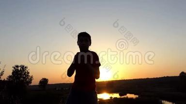 小男孩在秋天的日落时分在湖岸训练拳击