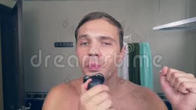 一个有趣的帅哥在浴室里用电动剃须<strong>刀刮胡子</strong>，在镜子前唱歌。 4k.