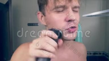 一个有趣的帅哥在浴室里用电动剃须刀刮胡子，在镜子前唱歌。 4k.
