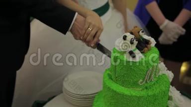 新娘和新郎在婚礼接待处切婚礼蛋糕。 切一个结婚蛋糕。 美丽的绿色节日蛋糕