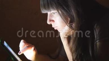 躺在沙发上有平板电脑的年轻女子。 女孩在家里使用平板电脑深夜。 在脸上闪耀着光芒