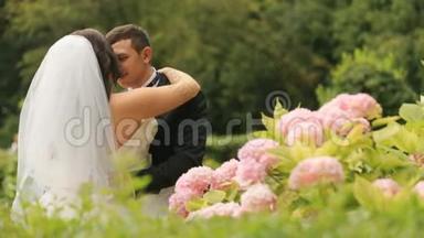 新婚新娘和新郎接吻快乐，在盛开的绿色夏日公园。 优雅的新郎拥抱华丽的新娘