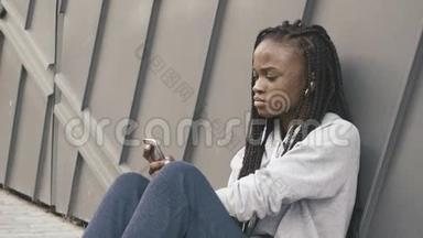 一位年轻美丽的非洲女孩坐在灰色纹理的墙边选择音乐倾听的肖像