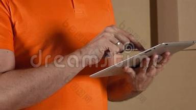 仓库工作人员穿着橙色制服，使用他的平板电脑与棕色纸箱堆叠的背景。 4K关闭视频
