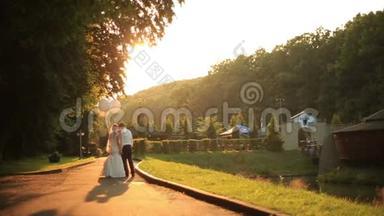 美丽的新婚夫妇带着气球在金色的秋天公园散步，在金色的夕阳下轻轻地接吻