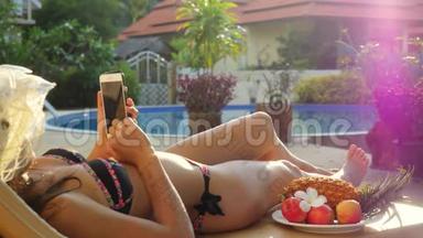 暑假，戴帽子的年轻女子躺在日光浴床上，在游泳池旁使用智能手机，带着美丽的风景