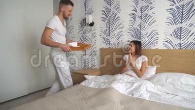 年轻人把咖啡放在床上，而他的女朋友穿着睡衣坐在床上