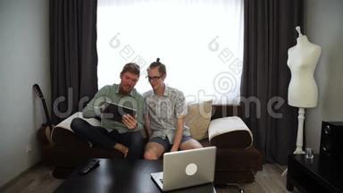 两个年轻人的兄弟正在家里工作，坐在沙发上，在他的平板电脑上，在台式笔记本电脑上工作。