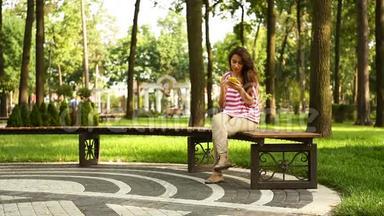 年轻的美女拿着<strong>手机</strong>坐在夏<strong>季</strong>公园的长椅上