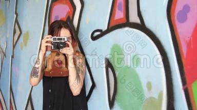 有纹身和老式相机的女孩站在涂鸦墙附近。