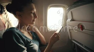日落时分坐在飞机窗口附近的旅游妇女在飞行中使用手机