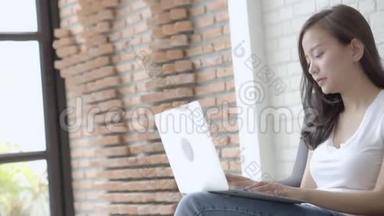 美丽的肖像亚洲年轻女子，微笑着在网上笔记本电脑工作，快乐地坐在客厅的沙发上